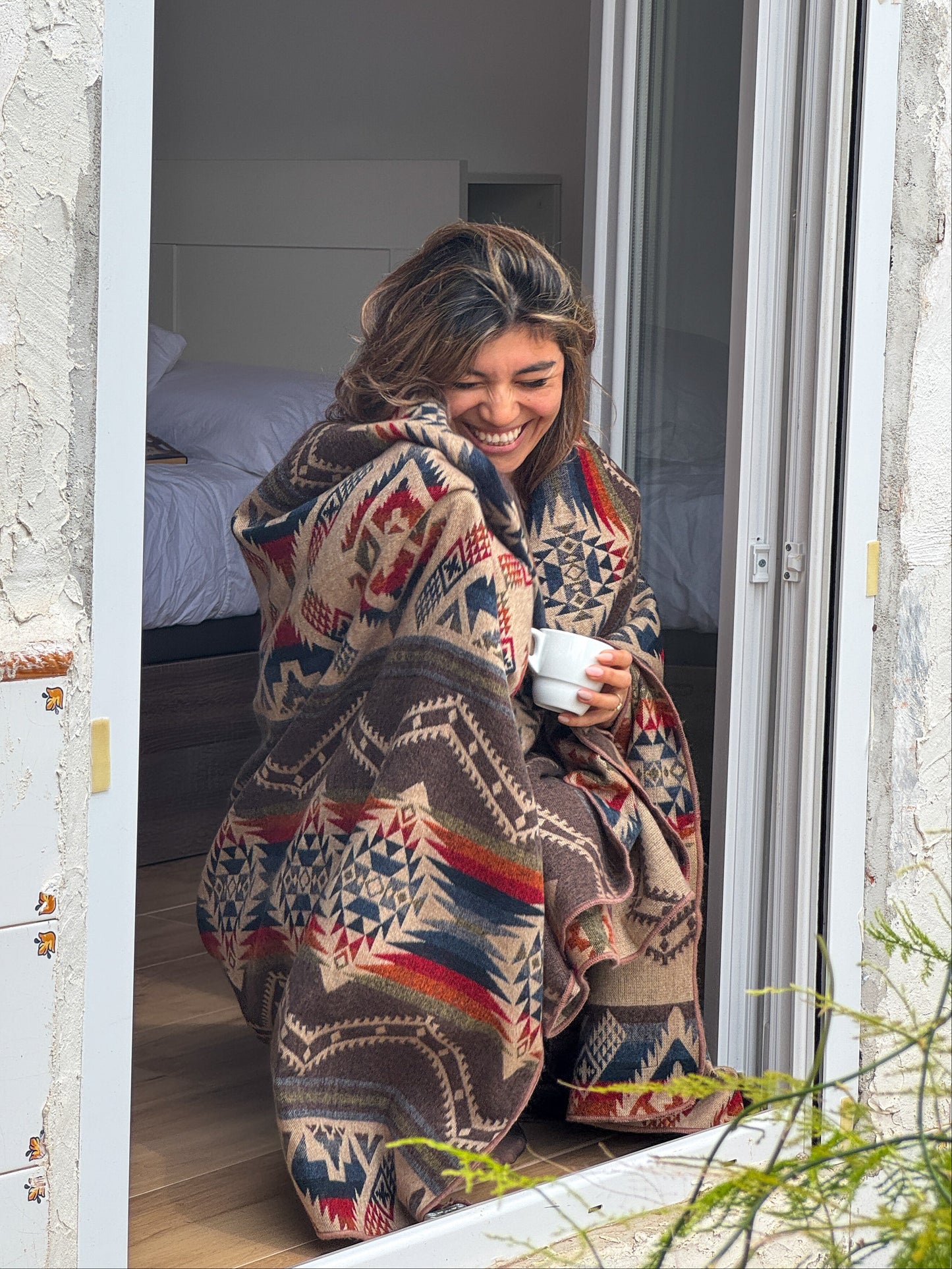 Woman wrapped in a patterned blanket, sitting on a windowsill holding a mug.