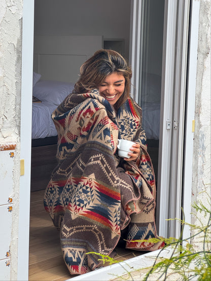 Woman wrapped in a patterned blanket, sitting on a windowsill holding a mug.