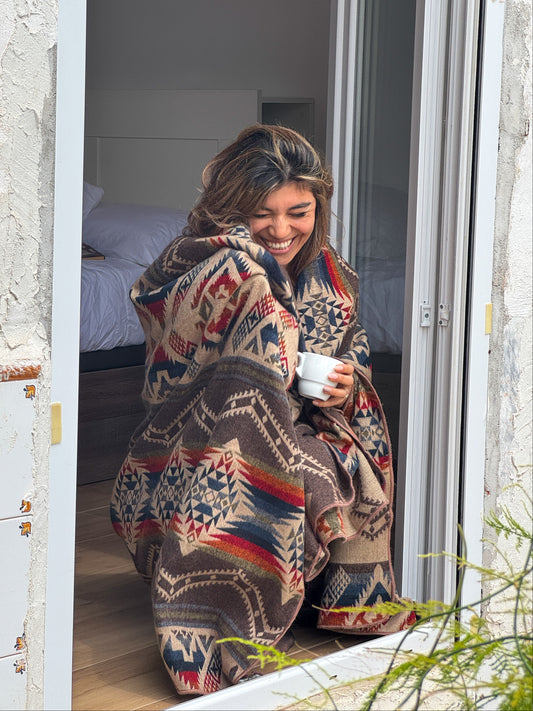 Woman wrapped in a patterned blanket, sitting on a windowsill holding a mug.